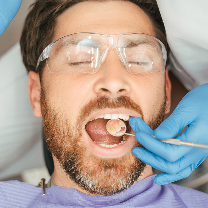 Attractive bearded man patient sitting in dental chair wearing protective glasses with open mouth